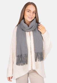 Femme portant une écharpe en laine grise à franges sur un cardigan crémeux tricoté et un pantalon beige, souriante, la main touchant l'écharpe.