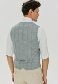 Męska niebieska kamizelka w kratę, wykonana z fakturowanej tkaniny, z tyłem na podwójne zapięcie z detalem w postaci klamry oraz białą koszulą z kołnierzykiem pod spodem.
