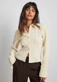 Camicia button-up in crema con texture e motivo ondulato, dotata di maniche lunghe e design cropped; abbinata a pantaloni marrone scuro.