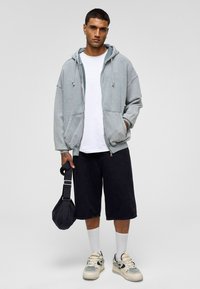 Grijze zip-up hoodie met kangoeroezakken, gecombineerd met een wit T-shirt en zwarte shorts. Het model draagt witte sokken en crèmekleurige sneakers en houdt een tas vast.