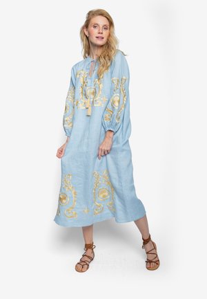 Jasnoniebieska lniana sukienka ze złotym haftem w kwiatowe wzory, szerokimi rękawami, dekoltem z paskiem do wiązania i luźnym krojem, zestawiona z brązowymi sandałami.