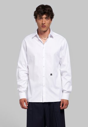 Jeune homme aux cheveux bouclés foncés portant une chemise blanche boutonnée et un pantalon foncé à fines rayures, debout devant un fond gris neutre.