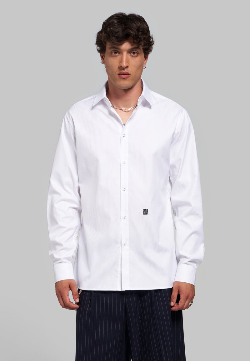 Jeune homme aux cheveux bouclés foncés portant une chemise blanche boutonnée et un pantalon foncé à fines rayures, debout devant un fond gris neutre.