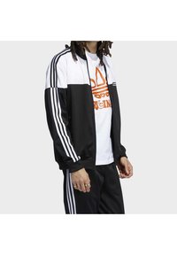 Svart och vit idrottsjacka med tre vita ränder på ärmarna, dragkedja, bärs över en vit t-shirt med en orange logotyp.