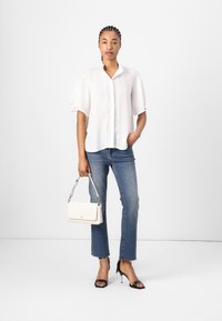 Blusa bianca con maniche a farfalla e bordo in pizzo, abbinata a jeans blu cropped con orli sfilacciati, sandali neri con tacco e una borsa bianca.