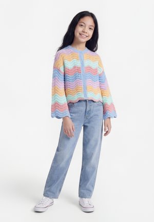 Dekle, ki stoji in se smeji, oblečena v pulover z rožnatim pastelnim mavričnim vzorcem s valovitimi robovi, svetlo modre kavbojke in bele superge na belem ozadju.