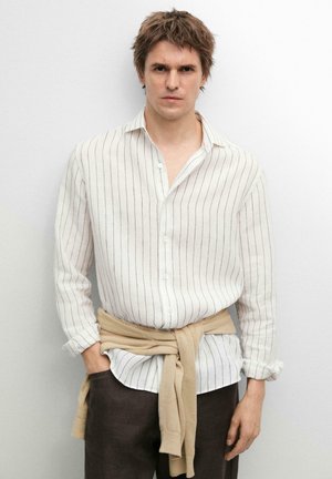 Mann trägt ein weißes, langärmeliges gestreiftes Hemd, einen beigefarbenen Pullover um die Taille gebunden und dunkle Hose, steht vor weißem Hintergrund.