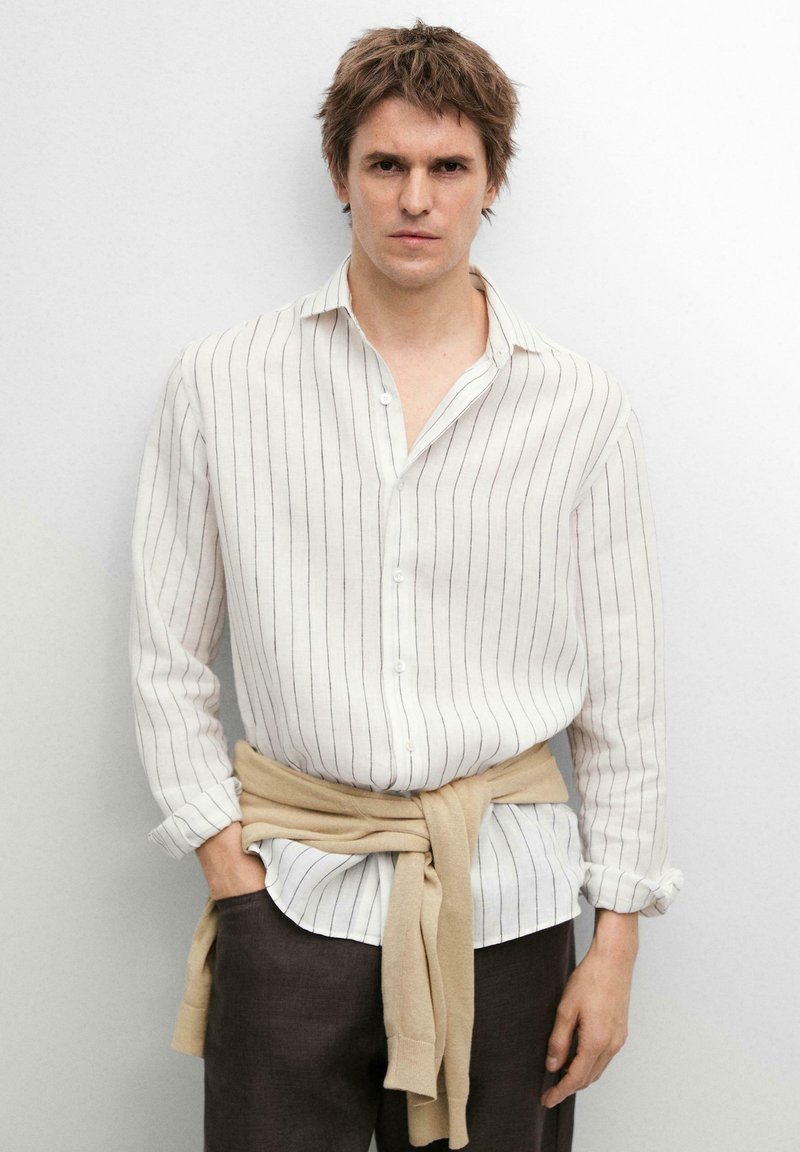 Man met een wit gestreept shirt met lange mouwen, een beige trui om de taille geknoopt en een donkere broek, staand tegen een effen witte achtergrond.