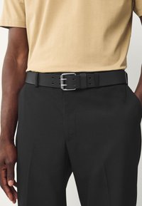 Black leather belt with a silver buckle, featuring adjustable holes, worn with beige t-shirt and black trousers. Smooth texture, simple design.