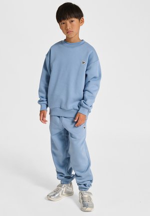 Hellblauer Pullover und passende Sweatpants aus weichem Stoff, mit einem kleinen Logodetail auf beiden Teilen, kombiniert mit silbernen Turnschuhen.