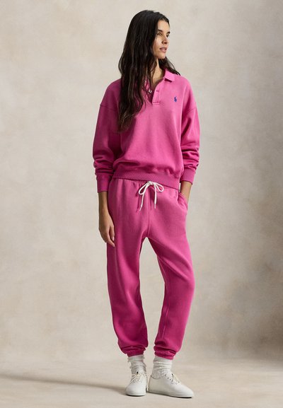 Φουξ φούτερ με γιακά πόλο και ριμπ μανσέτες, σετ με ταιριαστά παντελόνια φόρμας με ρυθμιζόμενο κορδόνι. Το λουκ ολοκληρώνεται με λευκά αθλητικά παπούτσια.