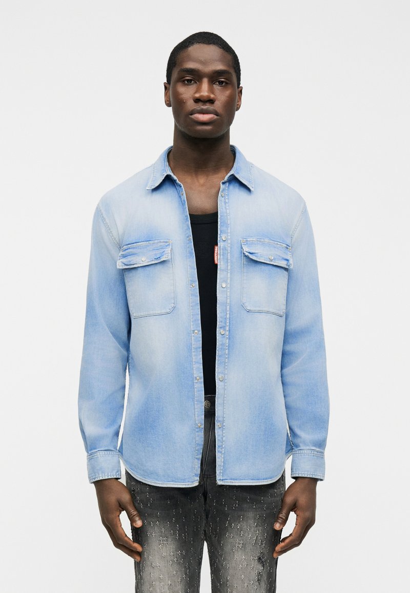 Άνδρας με σκουρόχρωμο δέρμα που φο ρά ανοιχτό γαλάζιο τζιν πουκάμισο πάνω από μαύρο τοπ και σκισμένο μαύρο τζιν, στέκεται μπροστά σε λευκό φόντο.