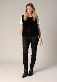 Zwarte nepbont vest over een witte shirt, gecombineerd met gestructureerde zwarte broek en enkellaarsjes. Heeft een slank ontwerp en een getailleerde silhouet.
