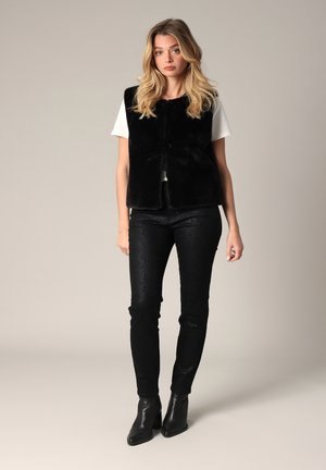 Gilet en fausse fourrure noire sur une chemise blanche, associé à un pantalon noir texturé et des bottines. Présente un design épuré et une silhouette ajustée.