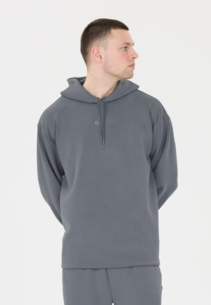 Jonge man in een grijze hoodie en bijpassende broek kijkt opzij met zijn handen achter zijn rug tegen een effen lichte achtergrond.
