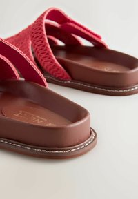 Sandales plates marron avec semelles épaisses et rembourrées, et larges brides en tissu rouge texturé sur le dessus.