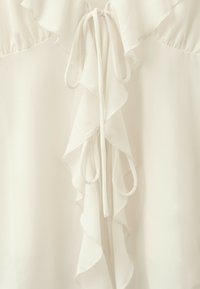 Blouse ivoire avec col à volants et détail de nouage devant, mettant en valeur la texture douce et fluide du tissu.