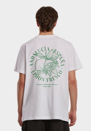 Person som har på seg en hvit T-skjorte med en illustrasjon av en grønn sitron og blader, og tekst på baksiden som lyder «Andalucía España Limón Fresco».