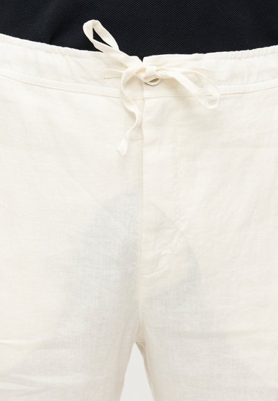 Λινά λευκά παντελόνια με μέση που έχει κορδόνι από ύφασμα δεμένο σε κόμπο, φορεμένα με μαύρο τοπ.