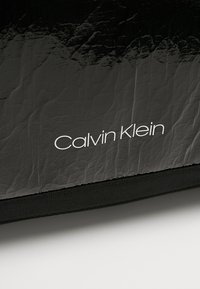 Czarna skórzana torba o pogniecionej teksturze z białym logo "Calvin Klein". Posiada trwały czarny materiał na dolnej części.