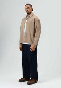 Homme aux cheveux tressés portant une chemise beige boutonnée, un sous-vêtement blanc, un pantalon sombre ample et des chaussures de randonnée marron, debout devant un fond gris.