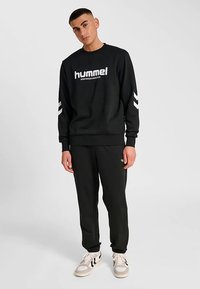 Svart sweatshirt med vitt "hummel"-logotyp och vita chevronränder på ärmarna, kombinerad med matchande svarta byxor och ljusfärgade sneakers.