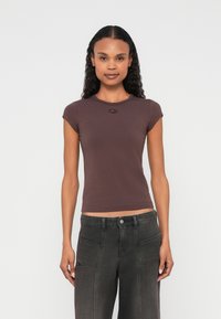 Žena s kučeravými vlasmi v priliehavom fialovom krátkom rukáve a tmavosivých širokých nohaviciach s vysokým pásom stojaca na bielom pozadí.