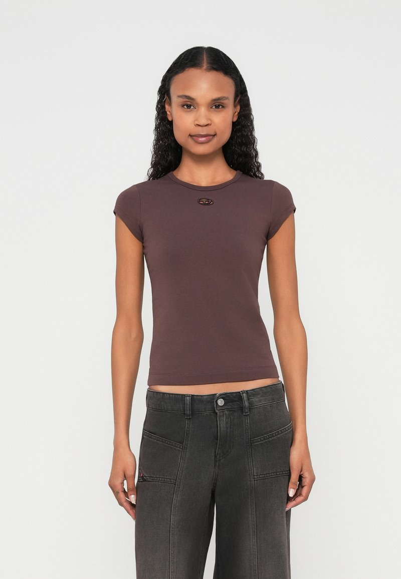 Žena s kučeravými vlasmi v priliehavom fialovom krátkom rukáve a tmavosivých širokých nohaviciach s vysokým pásom stojaca na bielom pozadí.