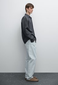 Casual outfit featuring a dark linen shirt, light blue relaxed-fit pants, and brown slip-on shoes, set against a minimalistic background.