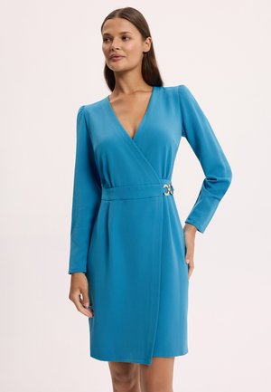 Kobieta ubrana w niebieską sukienkę kopertową z długim rękawem, dekoltem w kształcie litery V, talią podkreśloną oraz złotą klamrą, pozująca na tle jednolitego jasnego tła.
