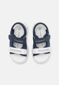 Granatowe sandały dla dzieci z białymi podeszwami, wyposażone w regulowane paski na rzep. Na paskach znajduje się napis "TOMMY" i "HILFIGER". Miękka tekstura.