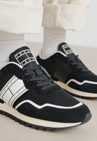 Zapatillas negras que combinan malla y ante, con acentos en blanco y un destacado logo de "TOMMY JEANS" en un diseño de cuadrícula. Suela de goma.