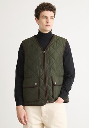 Quilted green vest with brown trim, featuring a full zipper, two snap-button pockets, and a structured design. Worn over a black turtleneck.