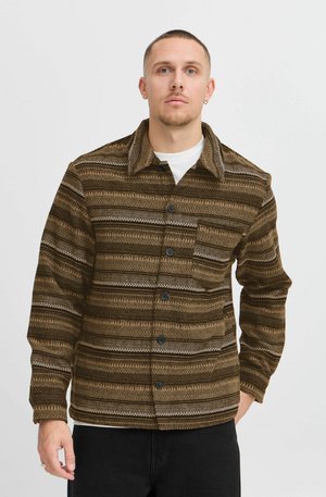 Hombre vestido con una chaqueta de botones marrón con estampado sobre una camisa blanca, de pie frente a un fondo blanco liso, mirando hacia adelante.