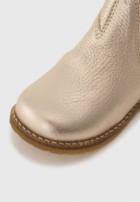 Gros plan sur la pointe d'une botte en cuir beige métallique avec une surface texturée, des coutures détaillées et une semelle en caoutchouc marron sur fond blanc.