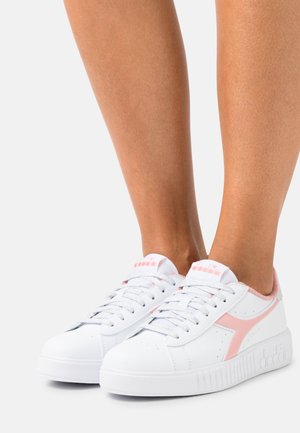 Baskets basses blanches avec des accents roses portées pieds nus, montrant les vues latérale et avant sur un fond uni clair.