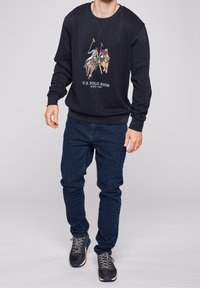 Marinblå sweatshirt med rund hals, prydd med en polo-inspirerad grafik och text. Tillsammans med mörkblå jeans och svarta träningsskor.