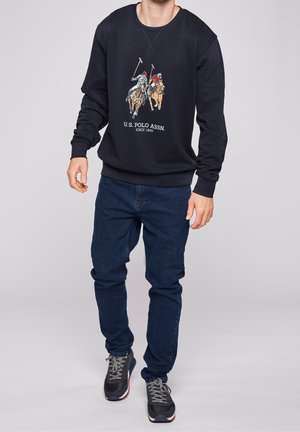 Sudadera azul marino con cuello redondo, con un gráfico y texto de temática de polo. Combinada con vaqueros azul oscuro y zapatillas deportivas negras.