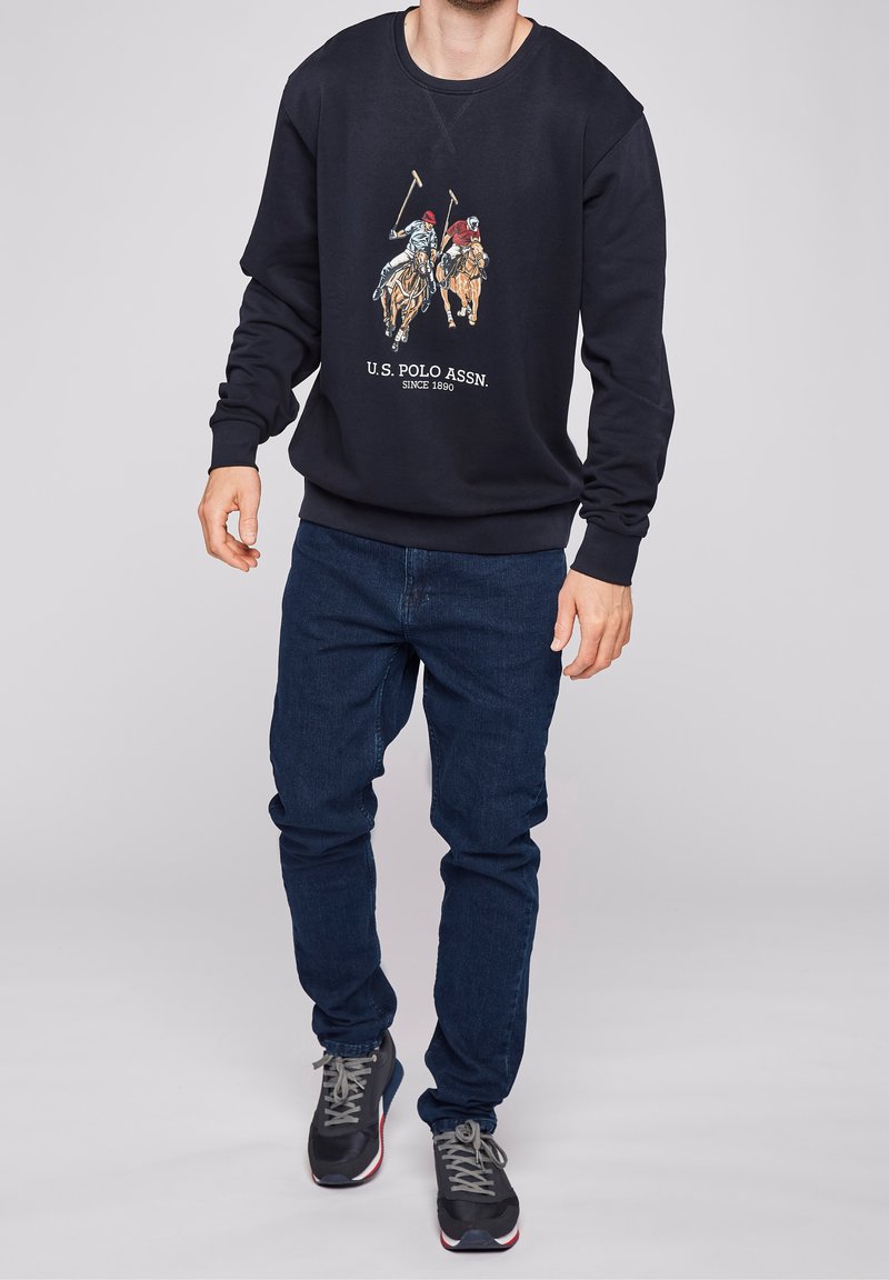 Marinblå sweatshirt med rund hals, prydd med en polo-inspirerad grafik och text. Tillsammans med mörkblå jeans och svarta träningsskor.