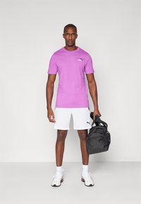 Lyslilla T-shirt, hvide shorts, sort gymnastikpose. Sneakers med hvide og sorte accenter. Modell står foran en simpel baggrund.
