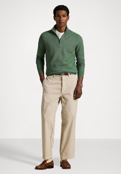 Green knit pullover with a zip neck, paired with beige trousers. Brown shoes and a watch complete the outfit, featuring a relaxed fit.