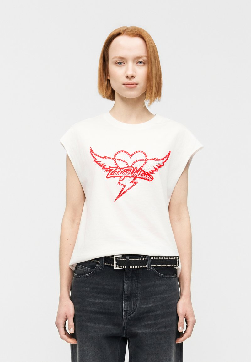 Λευκό βαμβακερό μπλουζάκι με κοντά μανίκια, με γραφικό κόκκινης καρδιάς με φτερά και το κείμενο "Zadig&Voltaire." Συνδυασμένο με σκούρο τζιν τζιν και ζώνη με καρφιά.