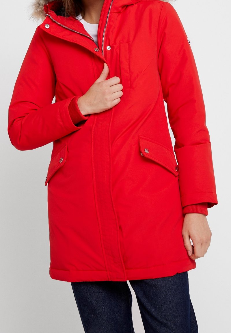 Rode gevoerde parka met ritssluiting en drukknopen, voorzien van een capuchon met nepbontvoering, voorzakken en geribbelde manchetten. Soepele textuur.