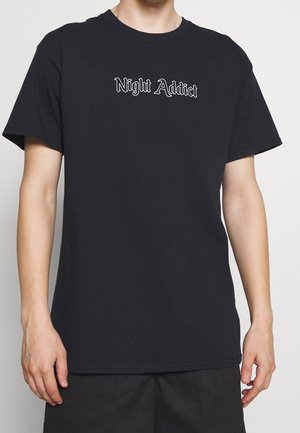 Osoba ubrana w czarny t-shirt z krótkim rękawem, na którym na środku klatki piersiowej znajduje się biały napis "Night Addict", na jednolitym tle.