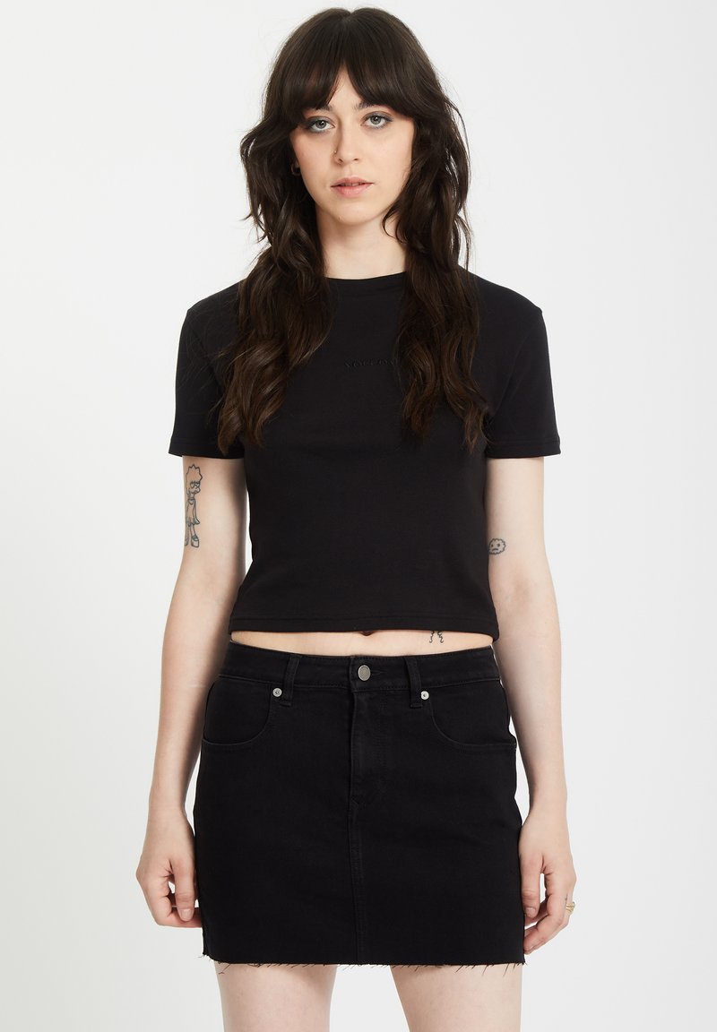 T-shirt noir court à manches courtes, arborant un logo discret au centre. Associé à une mini-jupe en jean noir avec un ourlet effiloché.
