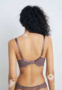 Soutien-gorge en dentelle mauve avec bretelles ajustables, design floral sur la bande, et fermeture à crochet à l'arrière. Tissu texturé avec un détail complexe.