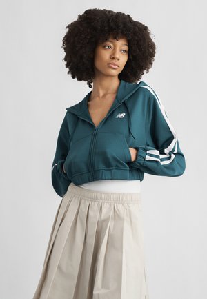 Mladá žena s kučeravými vlasmi, oblečená v tyrkysovej mikine na zips s bielymi pruhmi a béžovou plisovanou sukňou, ruky v vreckách, pozerá sa z boku.