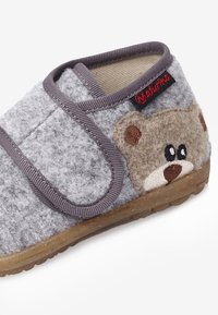 Szare filcowe kapcie z motywem misia, z zapięciem na rzep i brązowymi akcentami. Teksturowana podeszwa zapewnia przyczepność.