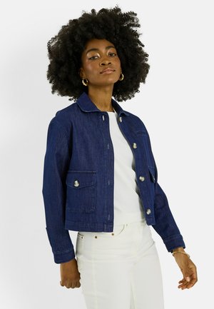 Femme aux cheveux bouclés portant des boucles d'oreilles créoles dorées, une veste en jean bleu foncé boutonnée, une chemise blanche et un pantalon blanc, debout sur un fond blanc.