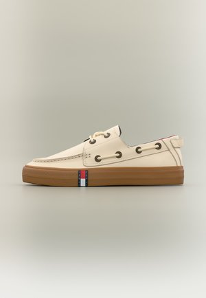 Zapato náutico de piel beige de caña baja con suela de goma marrón, detalles de cordones a través de ojales metálicos y una pequeña etiqueta con el logo en rojo, blanco y azul en la suela.